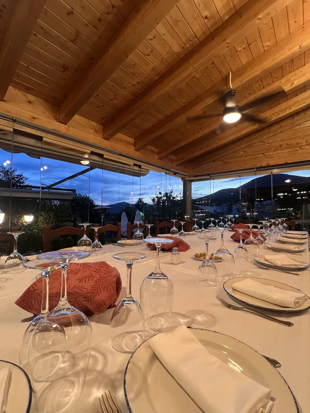 Mesa larga preparada para un grupo con las cristaleras abiertas y la Sierra de Guadarrama al anochecer